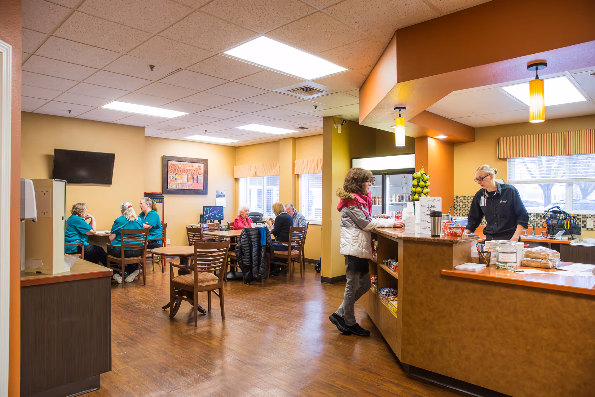 A communal dining area in a senior living facility with several people seated at tables conversing. A woman stands at a counter where a staff member is assisting her. The room has warm lighting, wood flooring, and large windows with blinds.