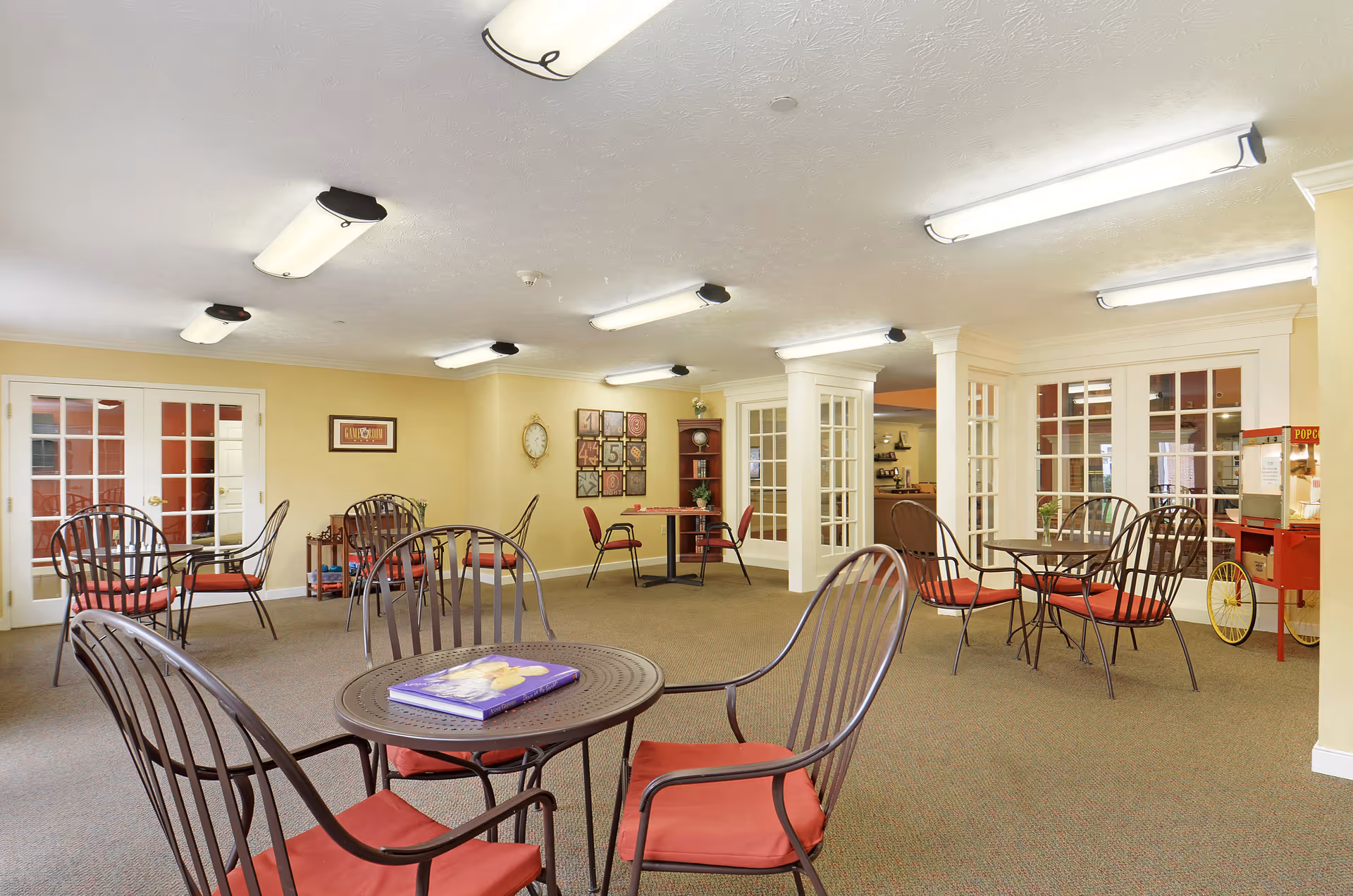 A spacious senior living facility common area with multiple round tables and metal chairs with red cushions. The room has beige walls, carpeted floor, and fluorescent ceiling lights. There are glass-paneled double doors and windows, a clock on the wall, decorative wall art, and a popcorn machine in the corner.