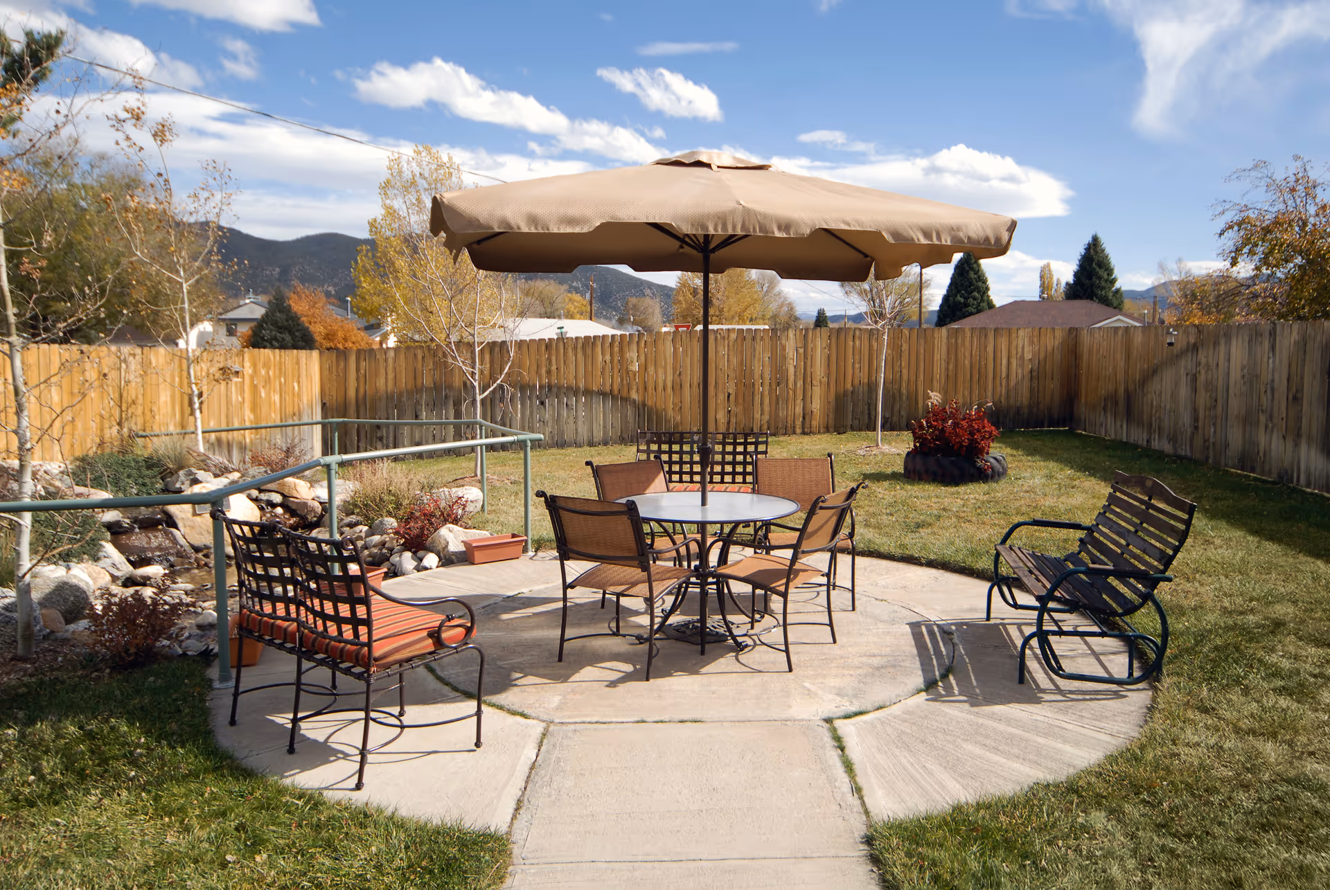 Fenced outdoor patio with a round table, chairs, and a large umbrella set on a concrete pad surrounded by grass and landscaping.