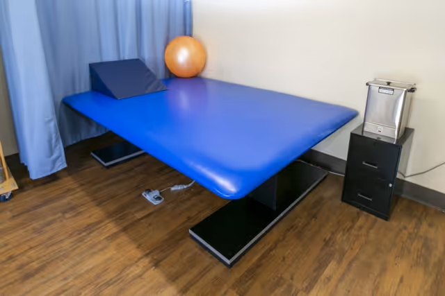 A blue padded treatment table in a therapy room with a wedge cushion, exercise ball, and small black drawers on a wood floor.