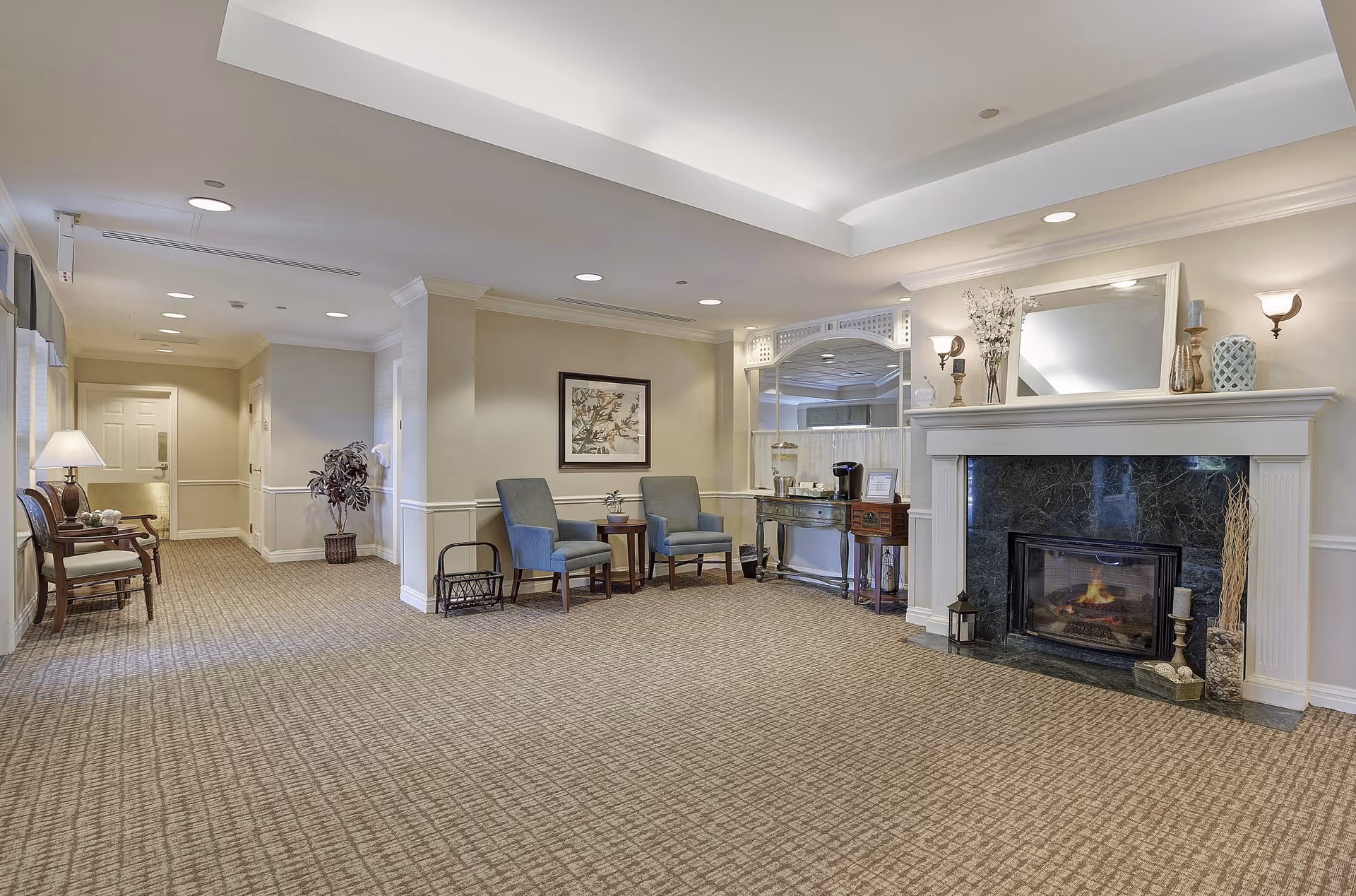 Bright common lounge in a senior living facility with chairs, side tables, and a fireplace under recessed ceiling lighting.