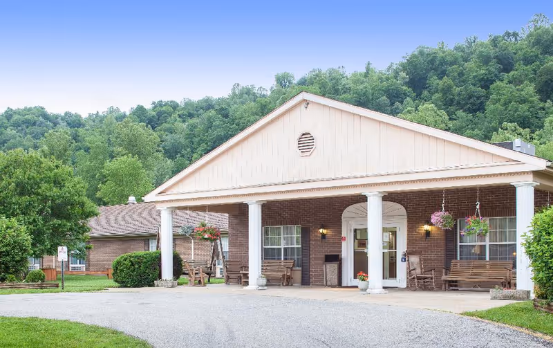 Front exterior view of Glenville Health and Rehabilitation building with a covered entrance supported by white columns, hanging flower baskets, wooden benches, and surrounded by green trees and bushes under a clear blue sky.
