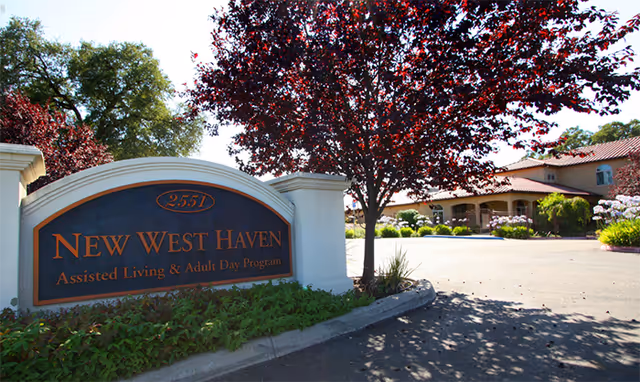 Entrance sign for New West Haven Assisted Living & Adult Day Program with a building and landscaped trees and bushes in the background under a clear sky.