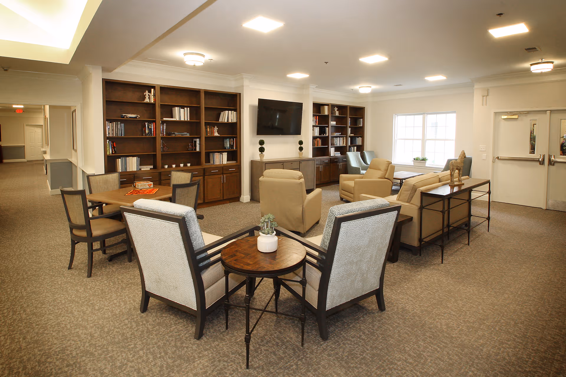 Well-lit communal lounge in a senior living facility with armchairs, side tables, built-in bookshelves, and a wall-mounted TV.