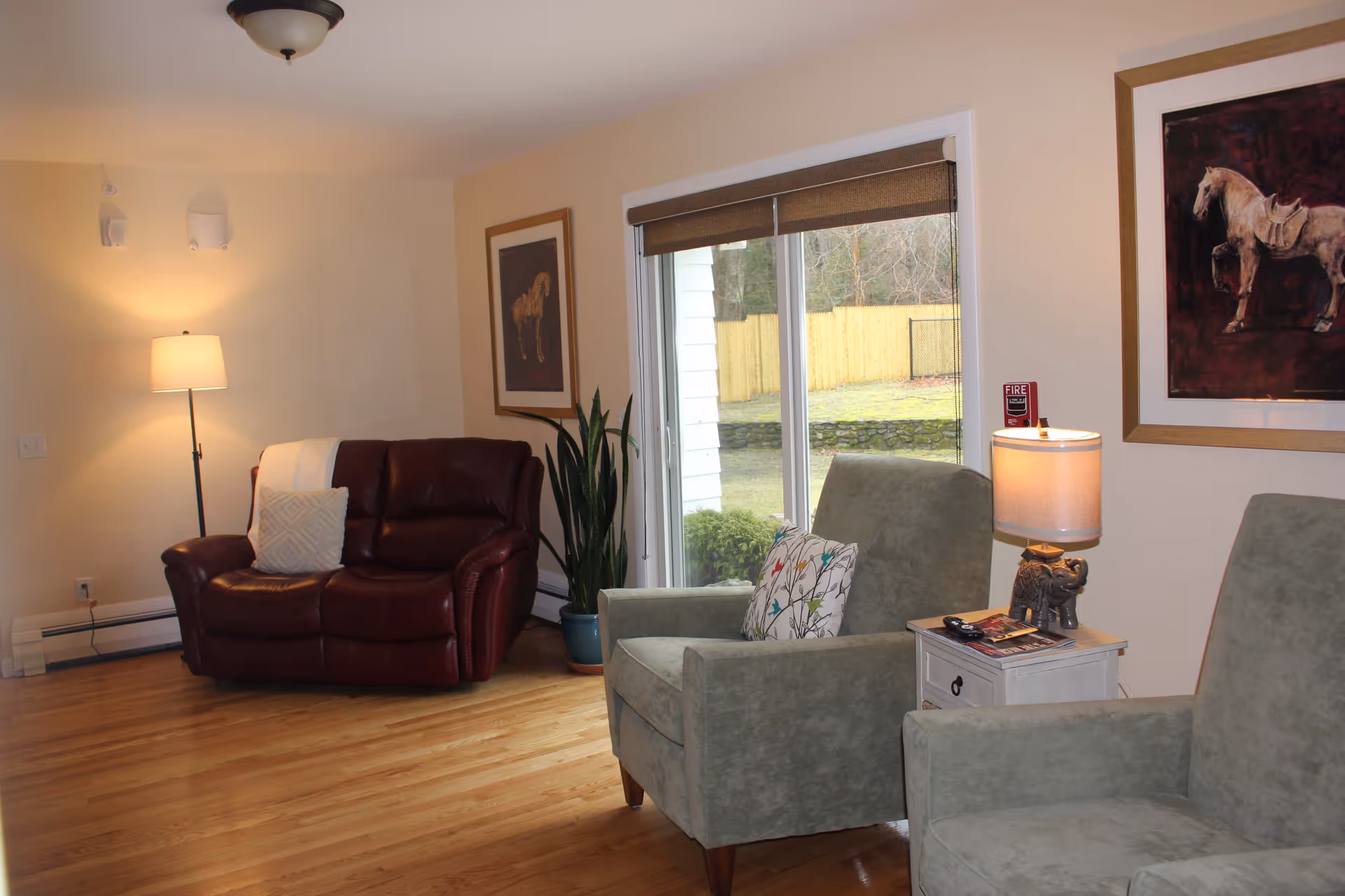 A cozy living room with hardwood floors, featuring a dark red leather loveseat with a white pillow and throw, two green upholstered armchairs with patterned pillows, a small white side table with a lamp and remote controls, a tall floor lamp, a potted plant, and two framed horse paintings on the beige walls. A large window with a brown roller shade shows an outdoor view with a wooden fence and trees.