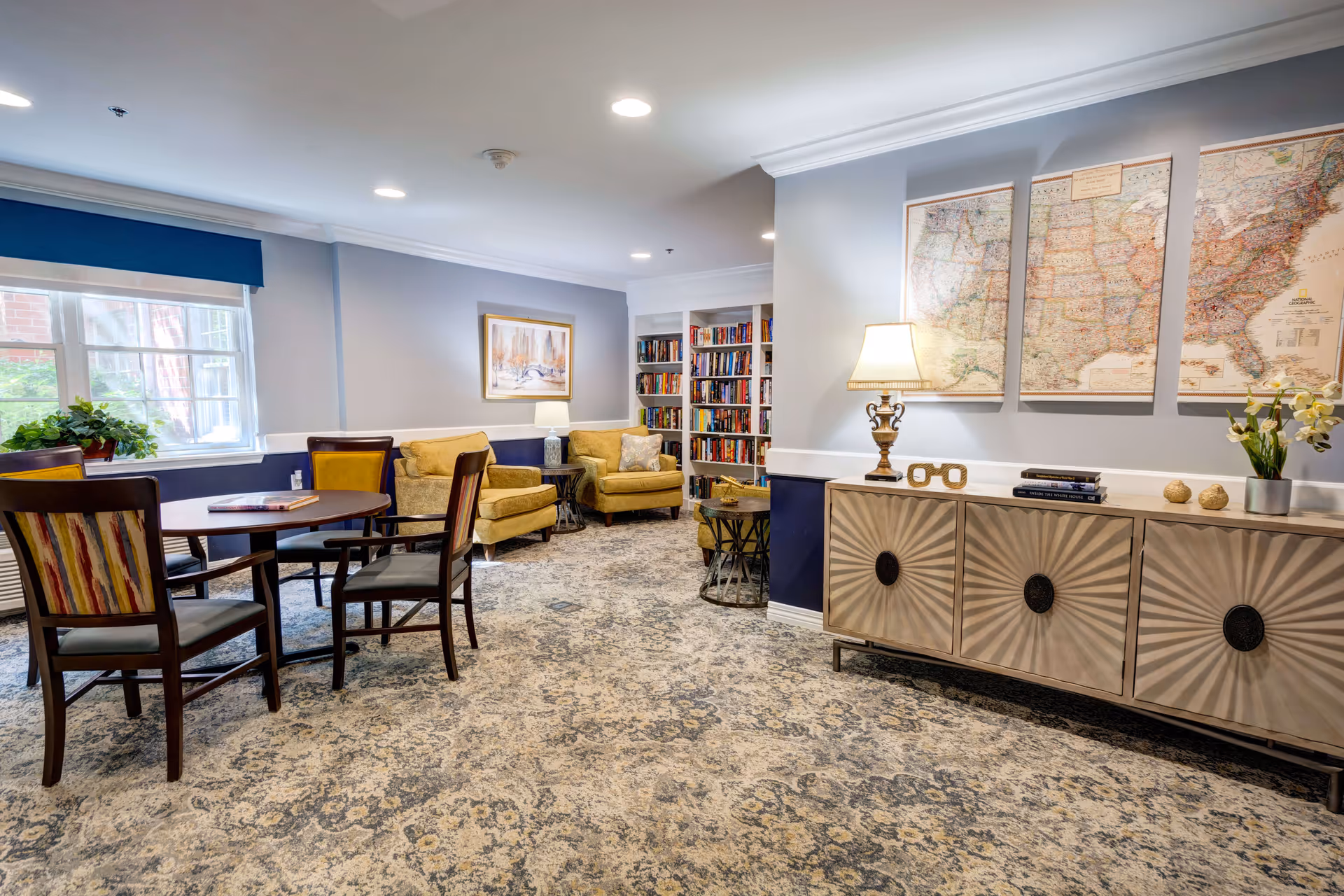 A cozy common area with a round table and four chairs on the left, two yellow armchairs with a small table and lamp between them in the back, a bookshelf filled with books, and a decorative sideboard with a lamp, books, and flowers on the right. Three framed maps hang above the sideboard. The room has patterned carpet and soft lighting.