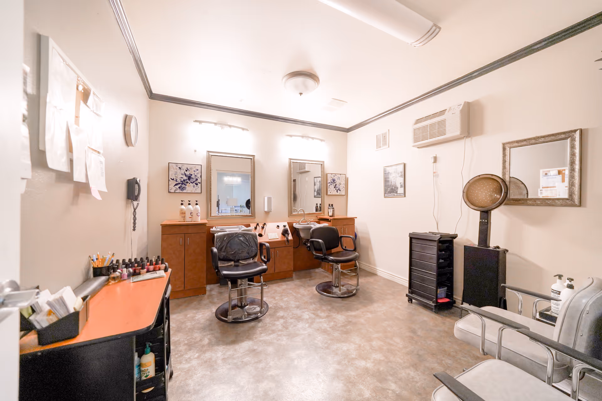 A small salon room with two black salon chairs in front of mirrors and wooden cabinets. The room has a hair dryer, nail polish bottles, and various salon supplies. The walls are light-colored with framed pictures and an air conditioning unit mounted on one wall.