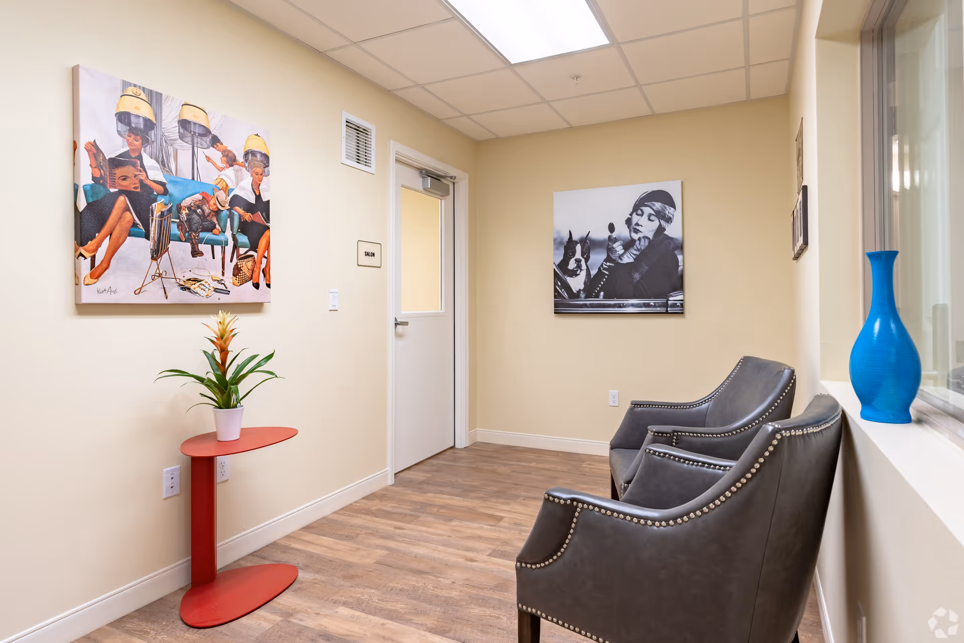 A waiting area with two gray armchairs with nailhead trim, a small red side table with a potted plant, and two framed pictures on the beige walls. One picture shows women under hair dryers, and the other is a black and white photo of a woman with a dog. There is a door labeled 'Salon' and a blue vase on the windowsill.