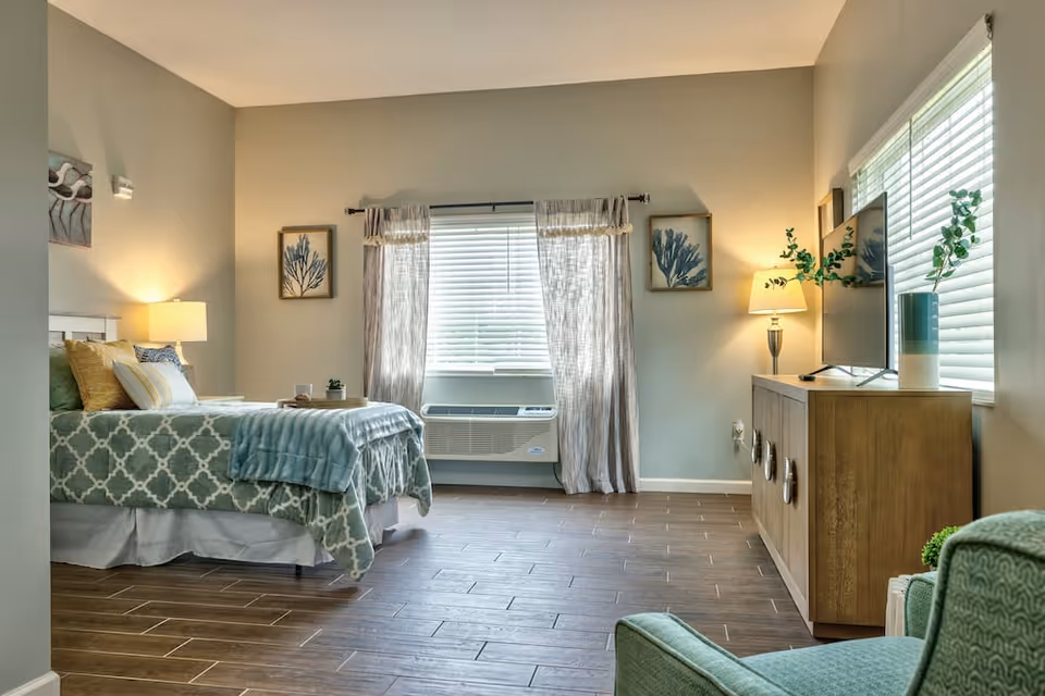 A well-lit bedroom with a bed on the left side covered with patterned bedding and multiple pillows. There are two framed artworks on the wall above the bed and a window with curtains and blinds in the center. Below the window is an air conditioning unit. On the right side, there is a wooden dresser with a lamp, a TV, and a vase with greenery. A green armchair is partially visible in the foreground on the right. The room has wooden flooring and soft, neutral-colored walls.