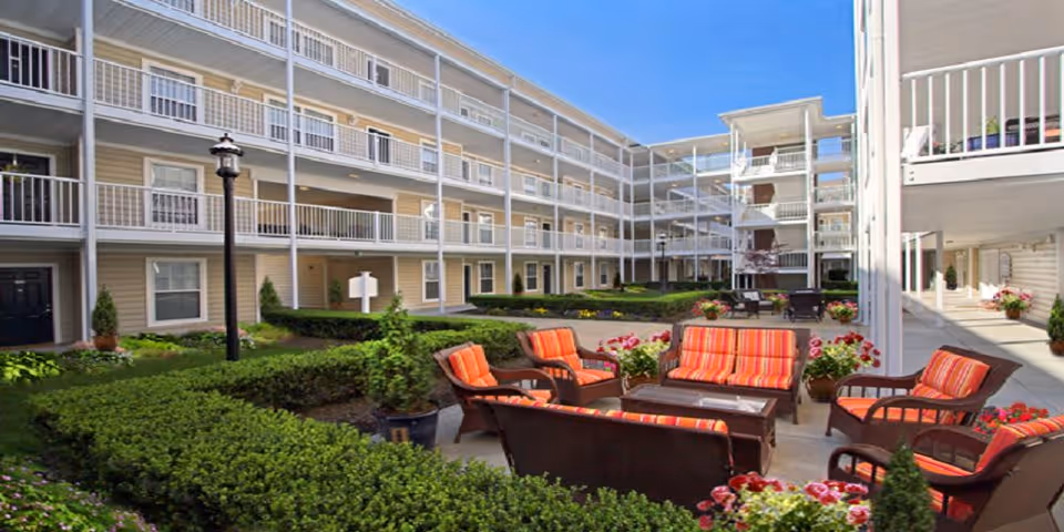 Outdoor courtyard area of The Commons at Centerbrooke Apartments featuring multiple seating arrangements with orange striped cushions, surrounded by green hedges and potted plants, enclosed by a multi-story apartment building with balconies and railings under a clear blue sky.