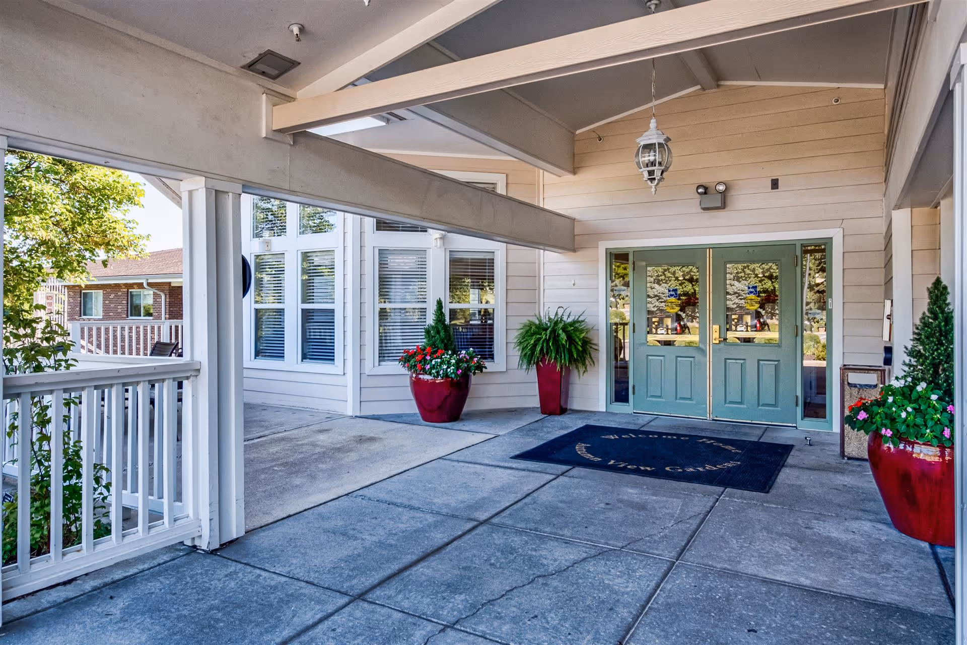 Covered entrance area of a building with double green doors, a welcome mat, and large red planters with green plants and flowers. The building exterior is light-colored with windows and a hanging lantern light fixture above the doors.