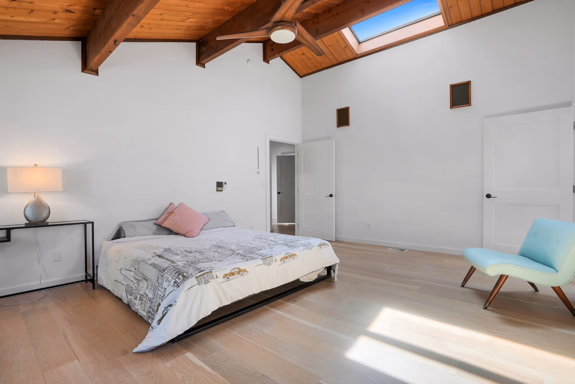 A spacious bedroom with a wooden ceiling and beams, featuring a bed with a cityscape-patterned duvet and a pink pillow. There is a modern lamp on a black metal side table to the left of the bed, a light blue chair with wooden legs on the right, and two white doors on the far wall. Natural light streams in through a skylight above.