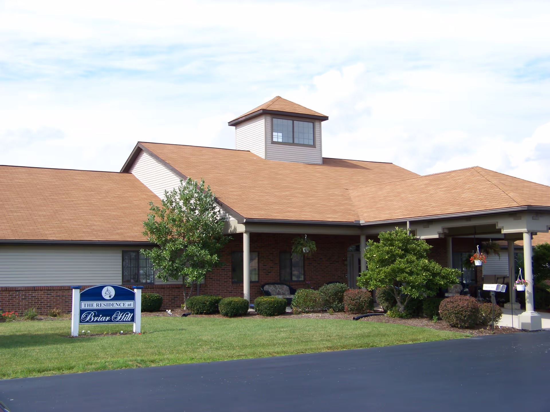 Exterior view of a single-story senior living facility building with a brown roof, beige siding, and brick accents. There is a covered entrance with columns, shrubs, small trees, and a sign on the lawn that reads 'The Residence at Briar Hill'.
