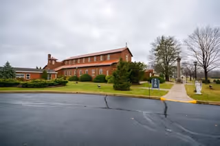 Exterior view of a large brick building with a cross on the roof, surrounded by green shrubs and trees on a cloudy day. A paved walkway leads to the entrance, and a sign is visible near the path.