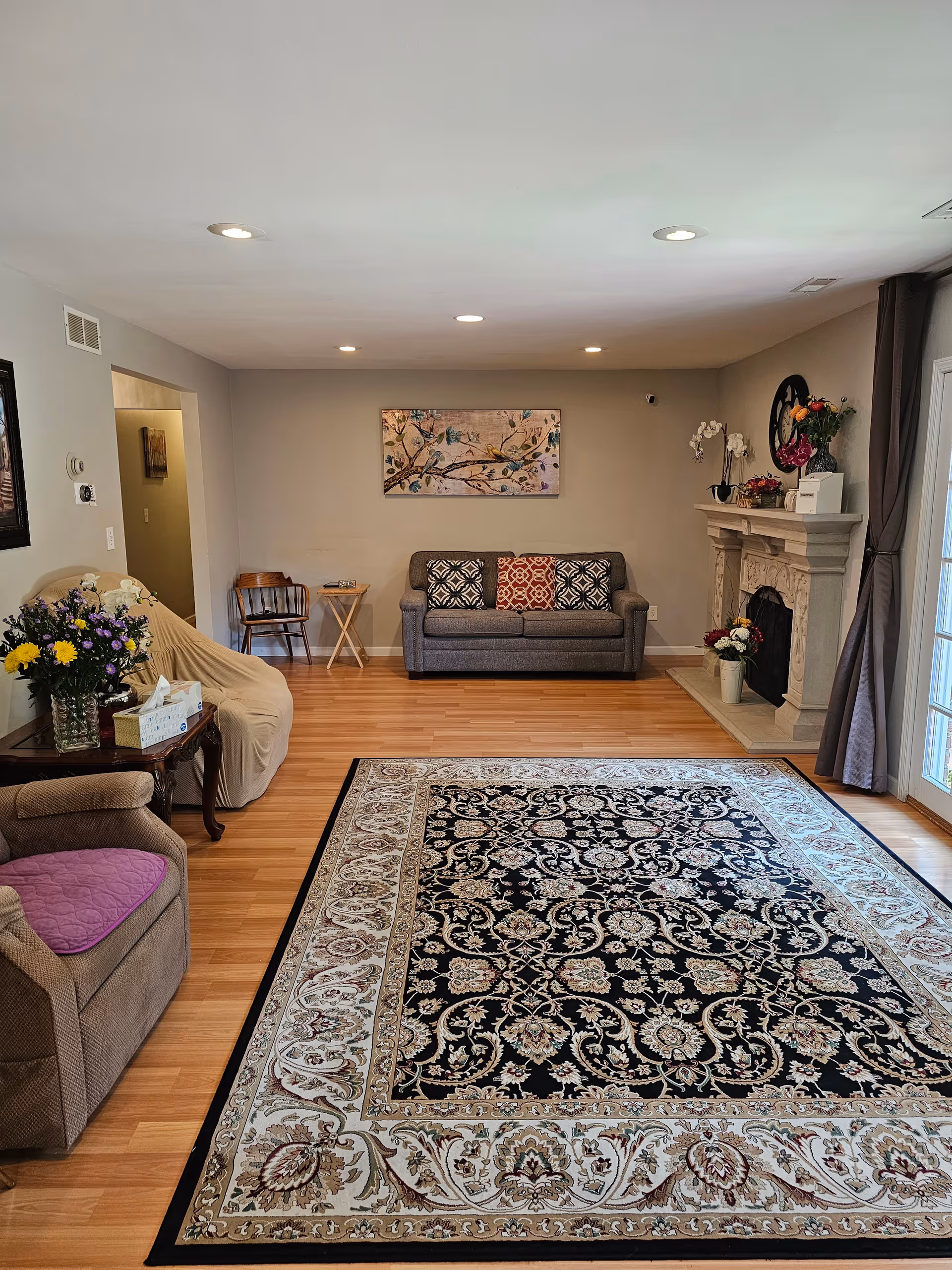 A cozy living room with wooden flooring, a large patterned area rug, a gray sofa with three decorative pillows against the far wall, a beige armchair with a purple cushion, a covered chair, a wooden side table with a tissue box and a vase of flowers, a fireplace decorated with flowers and a clock above it, and a painting of birds on a branch hanging on the wall.