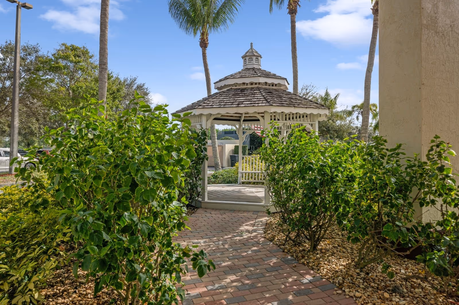 A paved walkway leads to a white wooden gazebo surrounded by green bushes and palm trees under a blue sky with some clouds.