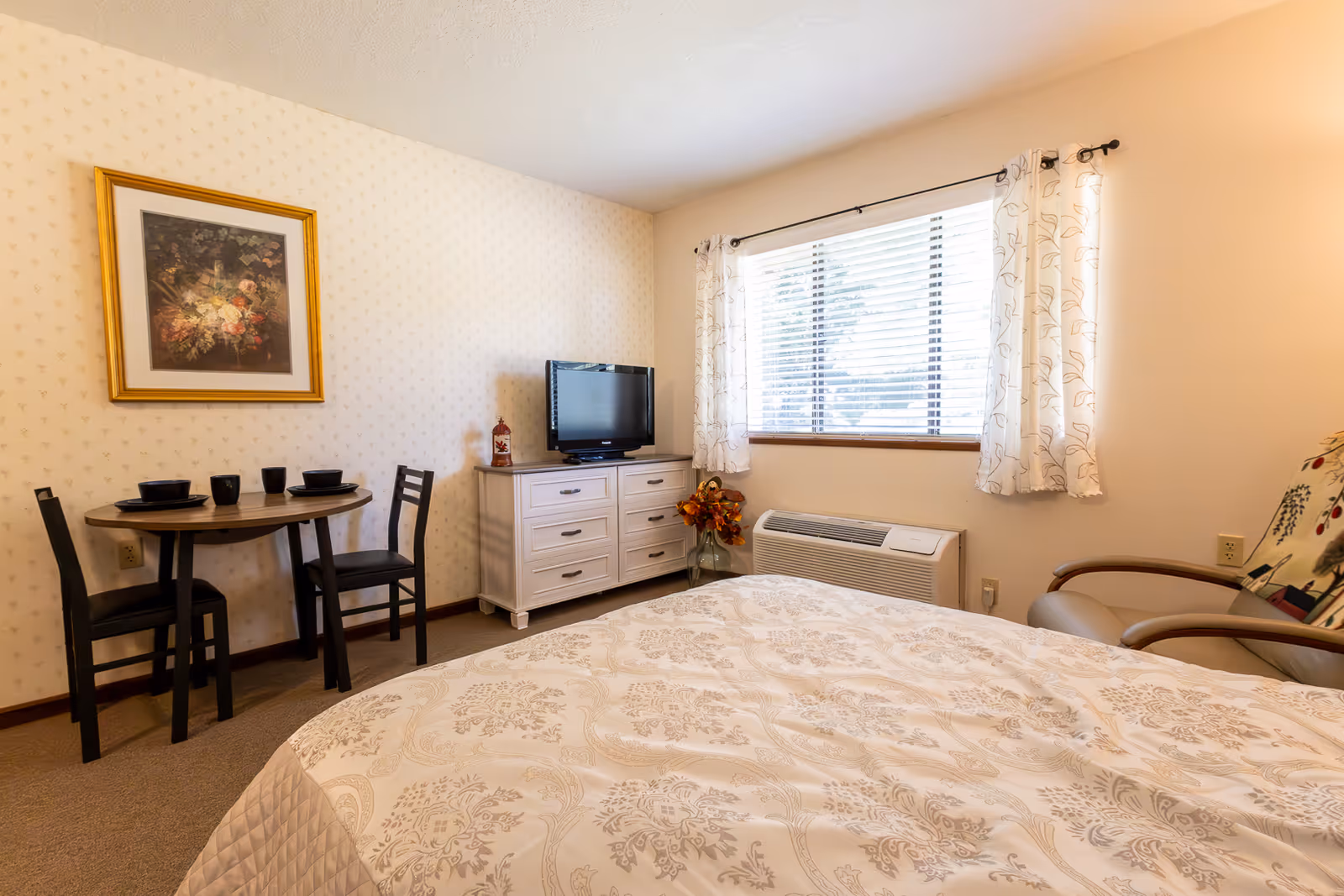 Well-lit furnished bedroom with a bed in the foreground, a small table and chairs, dresser with a TV, and a window with curtains.