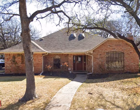 Exterior view of a single-story brick building with a walkway to the front door under trees.