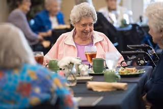 A group of elderly women sitting around a dining table, smiling and enjoying a meal together in a communal dining setting. The table is set with green cups, glasses of iced tea, plates of food, and a small floral centerpiece.