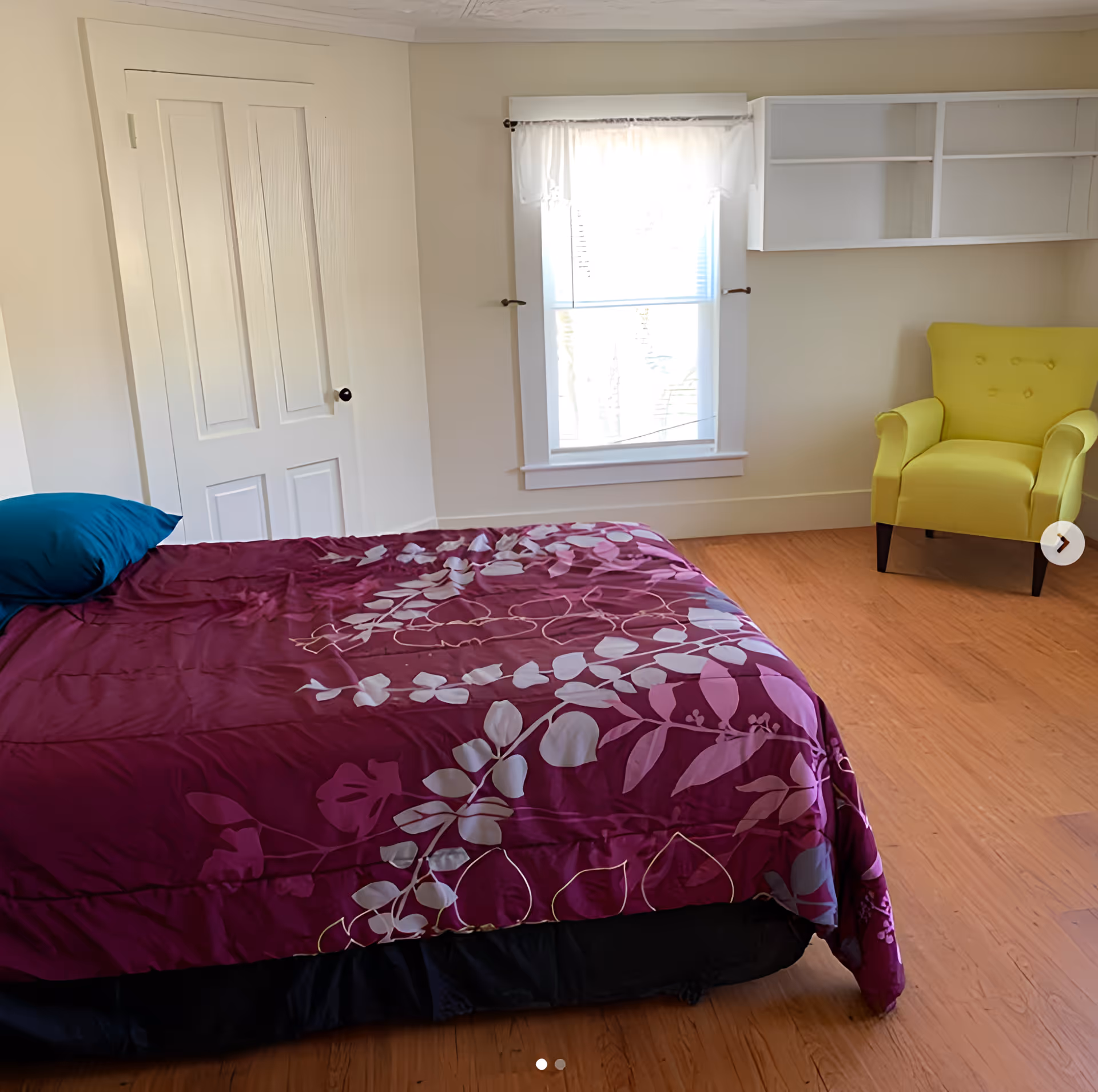 A bedroom with a bed covered in a purple floral bedspread and a teal pillow, a white door, a window with white curtains, a yellow armchair, and white wall-mounted shelves. The floor is wooden.