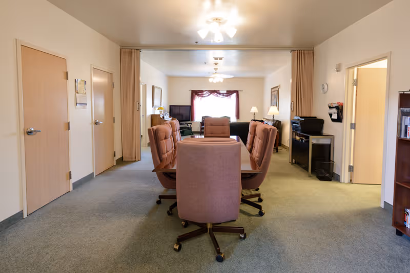 A well-lit interior room with a rectangular table surrounded by six cushioned office chairs on wheels. The room has beige walls, carpeted floor, and multiple wooden doors. In the background, there is a seating area with a TV, a window with a red curtain, and a floor lamp. The room appears to be a common or meeting area within a facility.