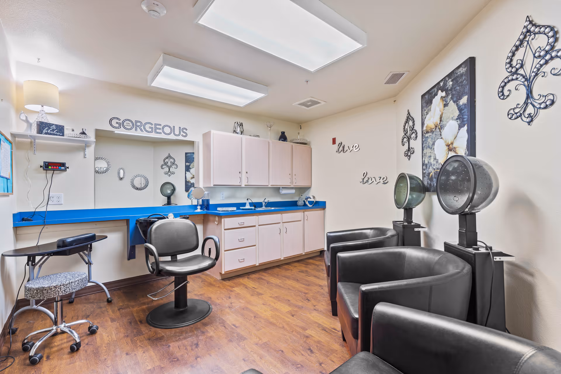 Interior view of a salon area in an assisted living facility with salon chairs, hair dryers, a large mirror with the word 'GORGEOUS' above it, cabinets, and wall decorations including the words 'live' and 'love'.