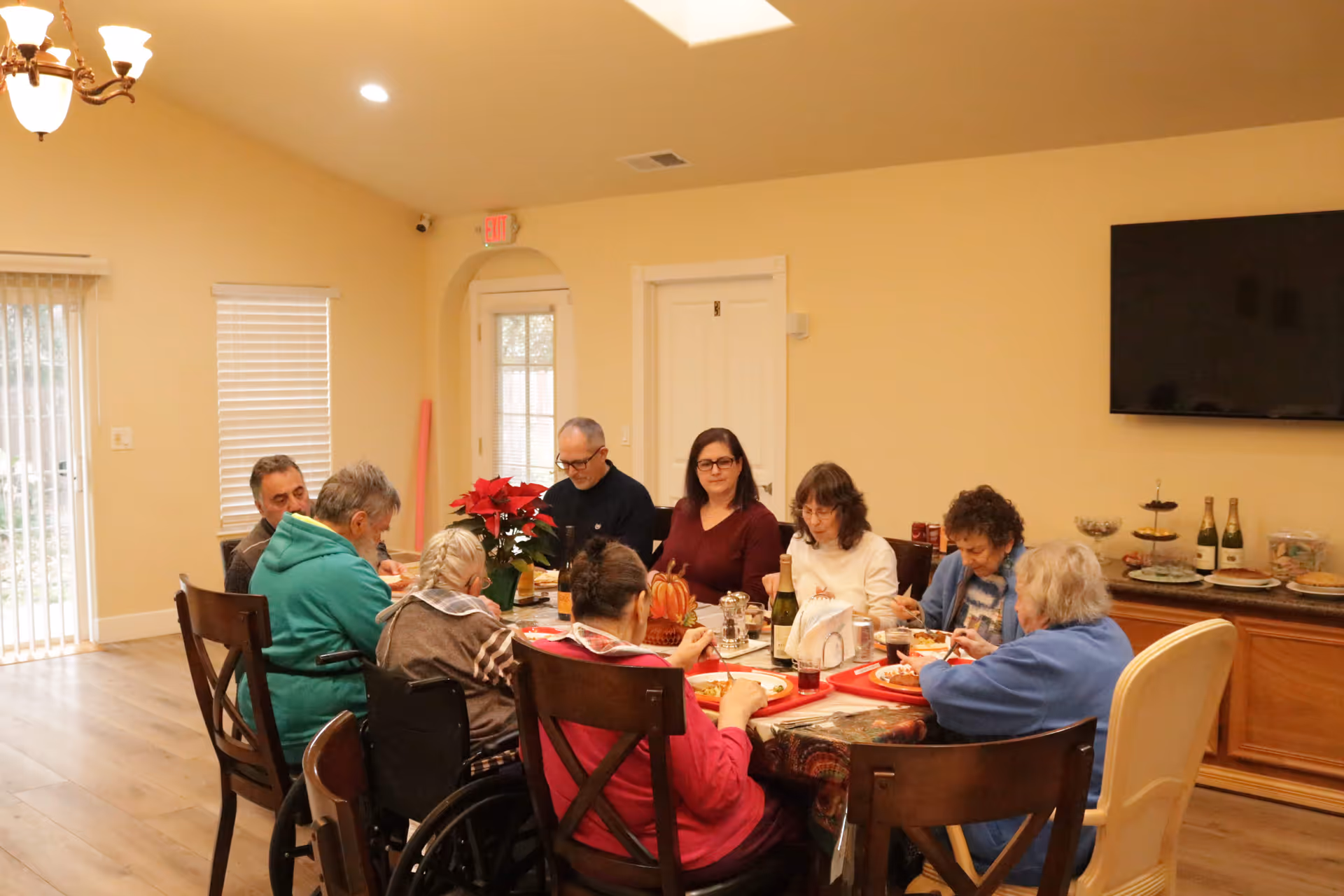 A group of elderly people and two caregivers sitting around a dining table in a well-lit room, sharing a meal together. The room has light-colored walls, a large flat-screen TV mounted on the wall, and a sideboard with bottles and plates. There is a poinsettia plant on the table, and the atmosphere appears warm and communal.