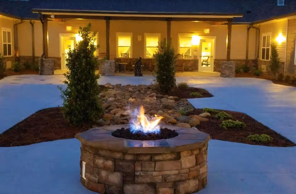 Evening courtyard showing a stone fire pit surrounded by landscaping with a lit building entrance in the background.