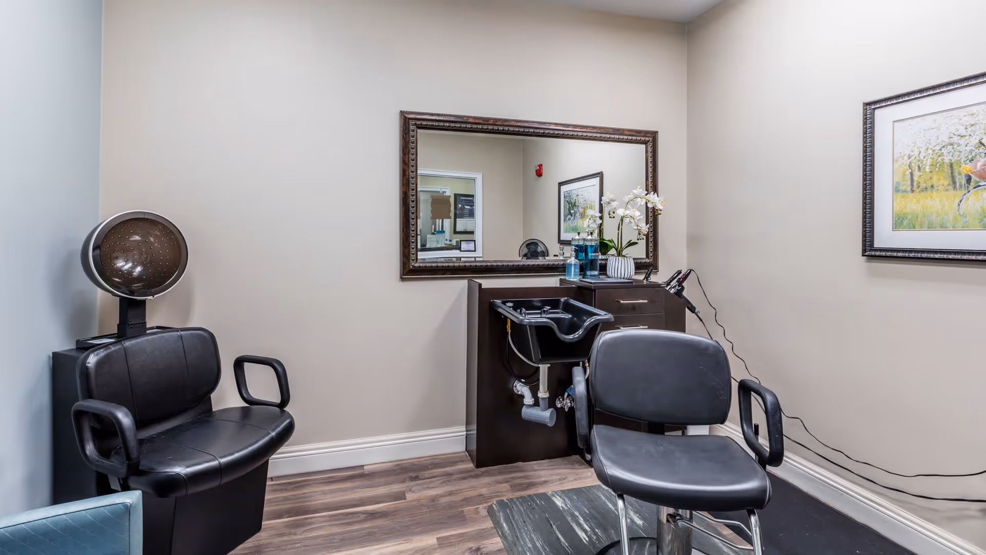 Interior of a small hair salon area with two black salon chairs, a hair dryer hood on the left, a black sink with a cabinet underneath, a large framed mirror on the wall, and a framed picture on the right wall. The floor is wooden and the walls are painted beige.