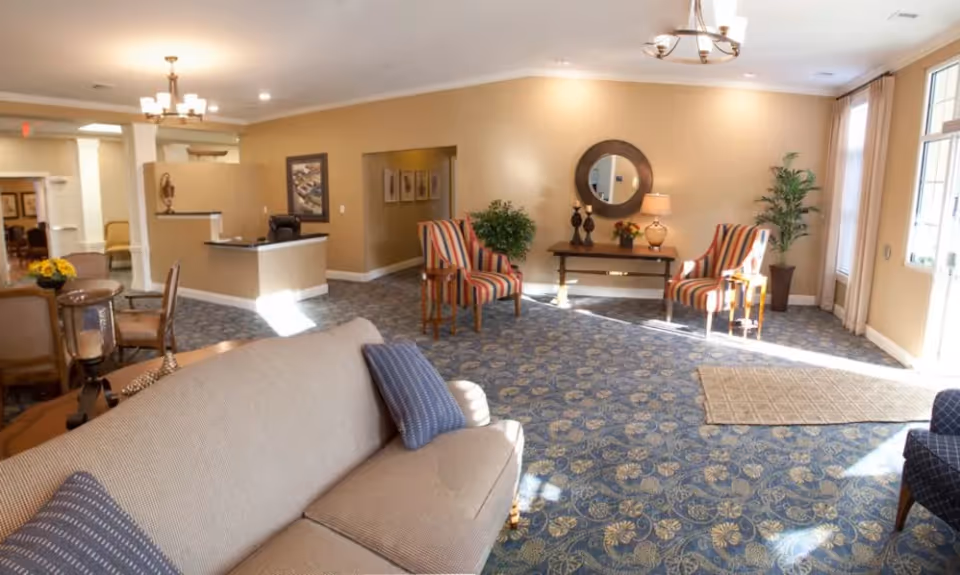 A spacious and well-lit senior living facility common area with beige walls and patterned blue carpet. The room features a beige sofa with blue pillows in the foreground, two striped armchairs near a wooden table with a round mirror and decorative items on the wall, and a reception desk in the background. Large windows allow natural light to fill the space, and there are plants placed around the room for decoration.
