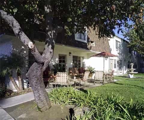 Outdoor patio area at a senior living facility with several cushioned chairs and a table under a red umbrella, surrounded by green grass, plants, and trees, adjacent to a two-story building with white and brown exterior walls.