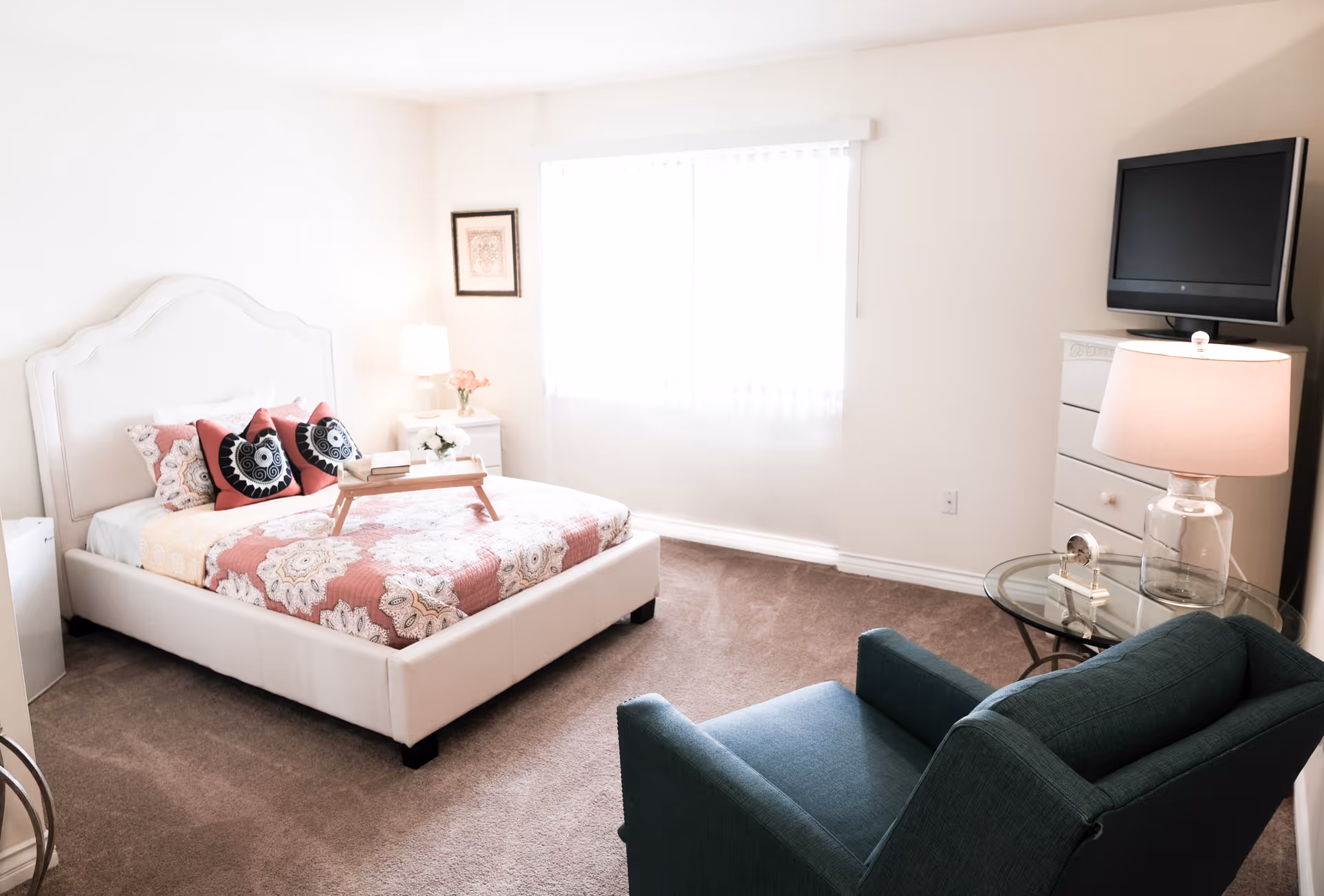Sunlit bedroom with a white upholstered bed dressed in patterned linens, a teal armchair, dresser with a TV and lamps.