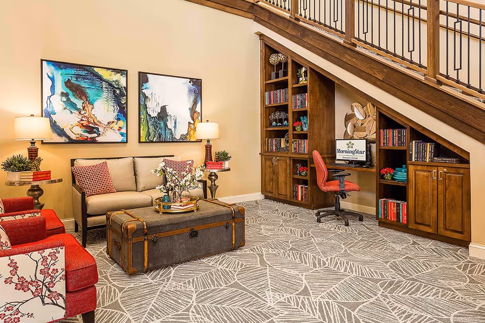 Comfortable sitting area with a sofa, red armchairs, a trunk coffee table, abstract wall art, and built-in bookshelves and desk under a staircase.