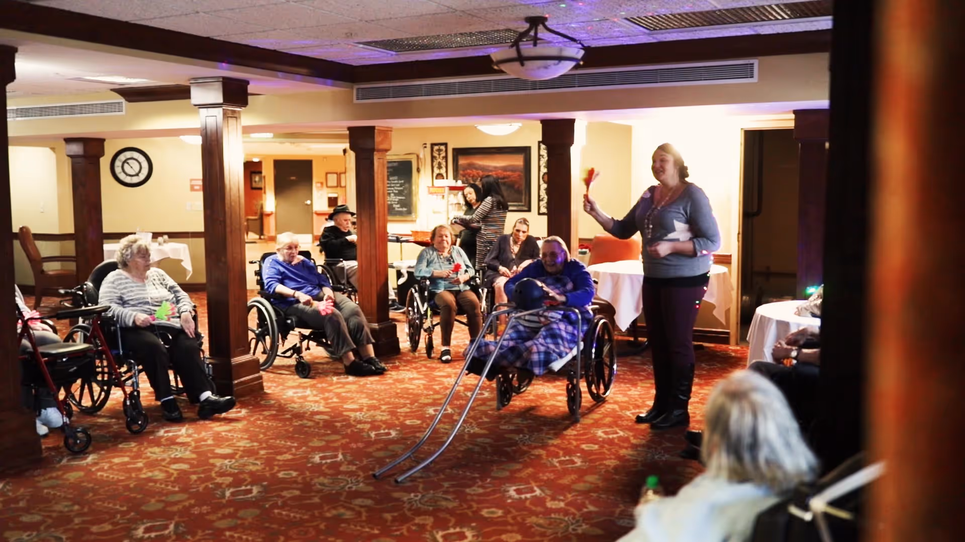 A group of elderly residents in wheelchairs taking part in a seated activity led by a staff member in a carpeted common room.