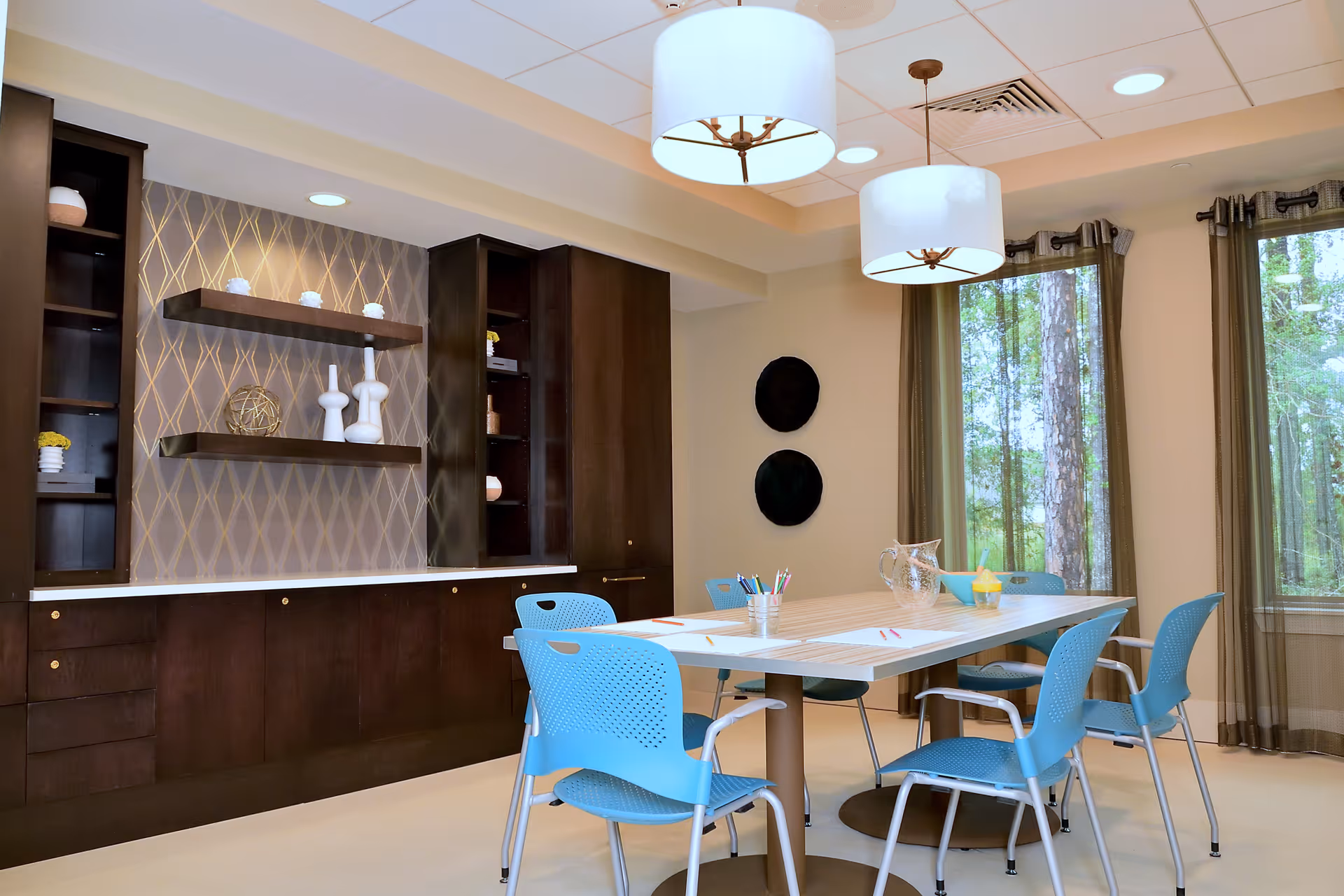 A bright and modern room with a rectangular table surrounded by six blue chairs. The table has some papers, colored pencils, a glass pitcher, and a bowl with a yellow object. The room features dark wood built-in cabinets and shelves with decorative items, patterned wallpaper, two large windows with sheer curtains showing a wooded outdoor view, and two large white pendant lights hanging from the ceiling.