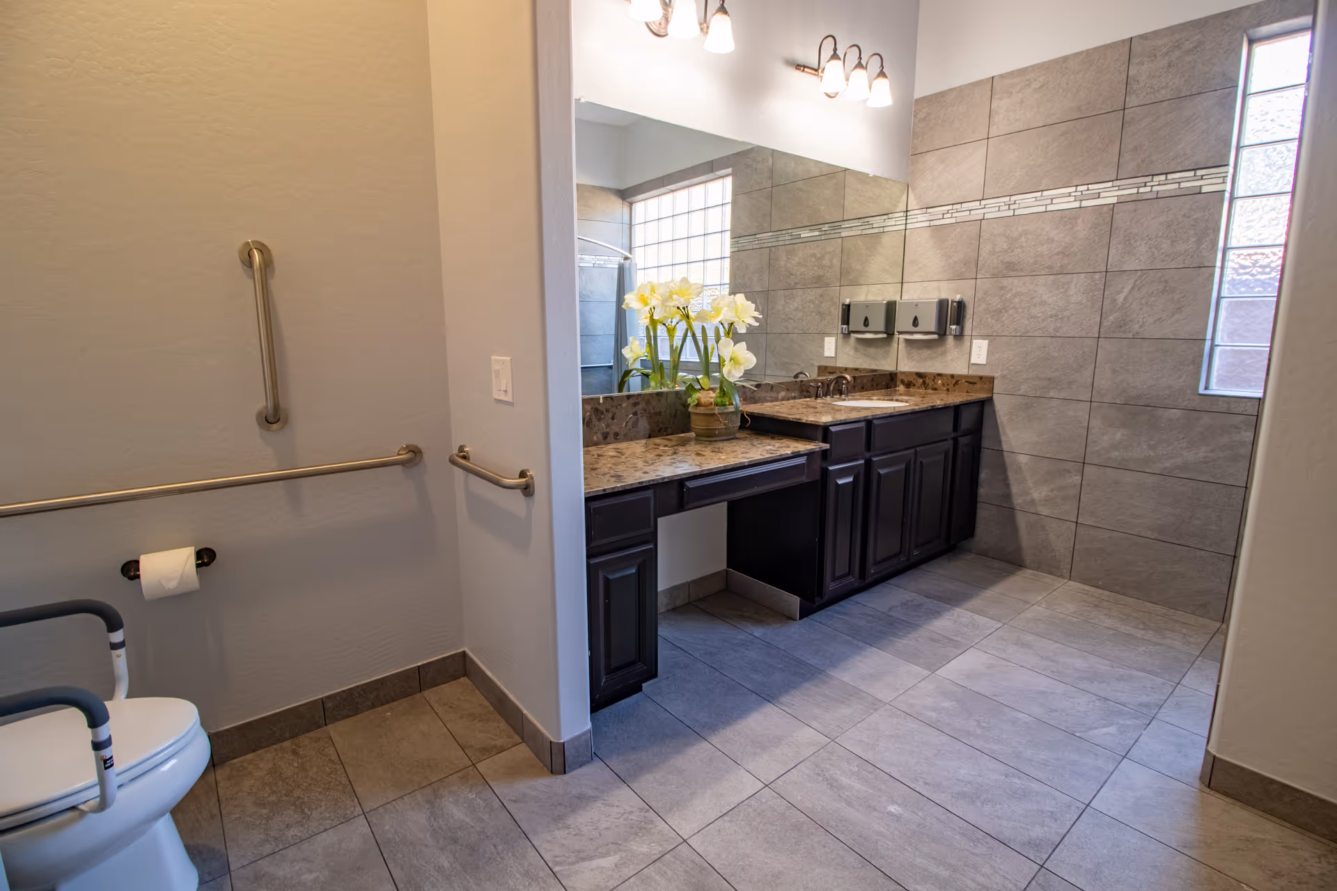 Spacious accessible bathroom with tiled floors, dual-sink vanity and large mirror, and a toilet with grab bars.