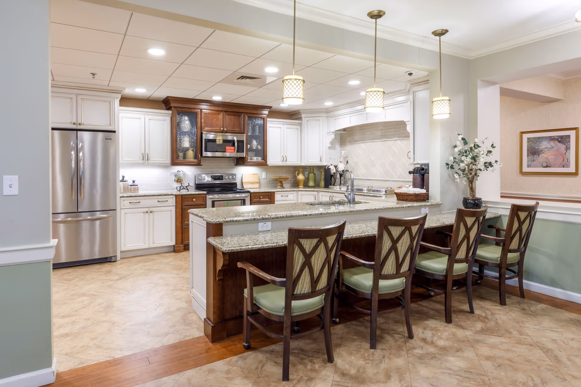 A spacious kitchen area with a large granite countertop island featuring five wooden chairs with green cushions. The kitchen has white and dark wood cabinetry, stainless steel refrigerator, oven, and microwave. Three pendant lights hang above the island, and there is a decorative plant on the right side near an open wall with a framed painting.