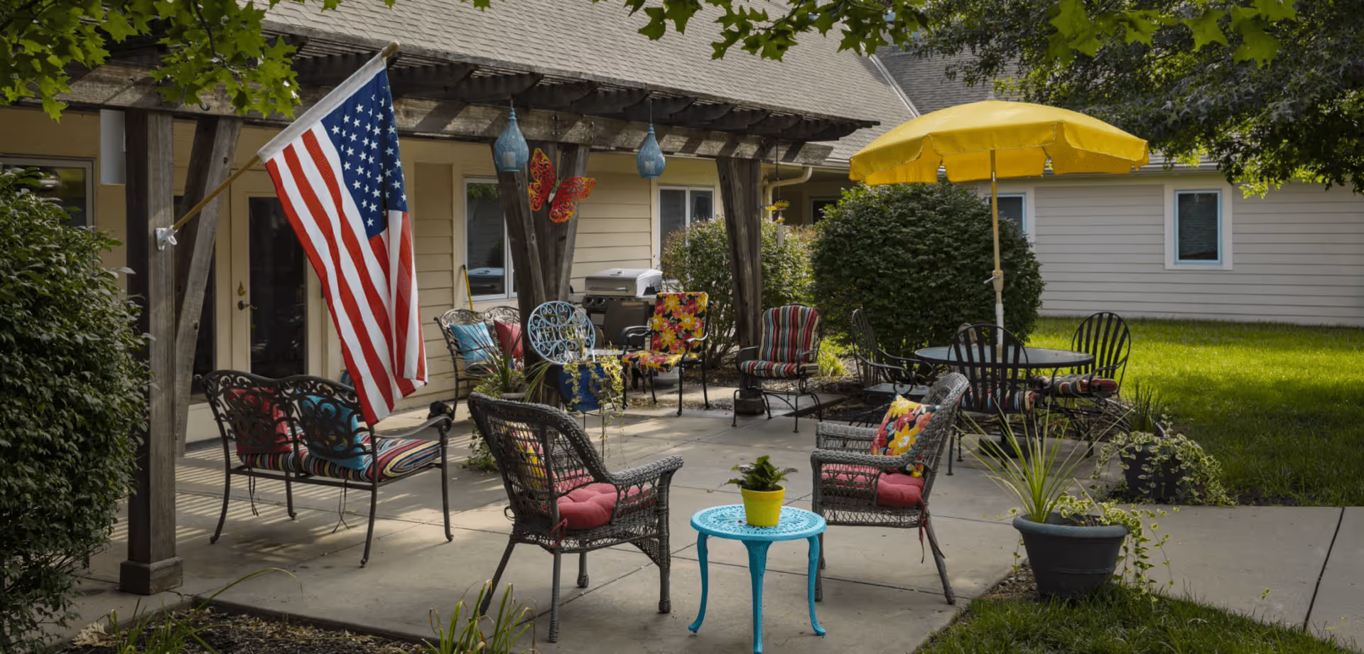 Outdoor patio area at Homestead Assisted Living of Shawnee with various chairs, a small blue table with a yellow potted plant, a yellow umbrella over a round table, and an American flag hanging from a wooden pergola. The area is surrounded by greenery and bushes.