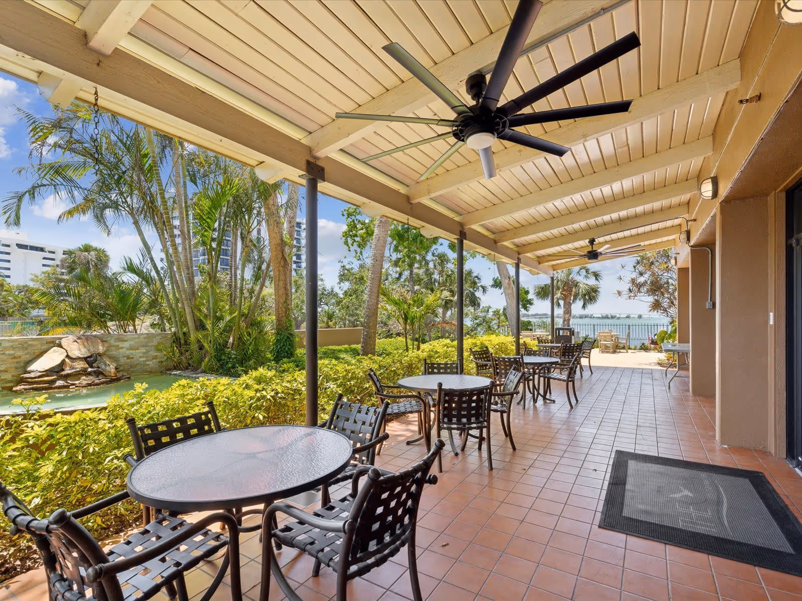 Covered outdoor patio area with multiple round glass tables and black metal chairs. The patio has a tiled floor, ceiling fans, and overlooks a garden with palm trees and a small water feature. In the background, there is a view of water and some buildings.