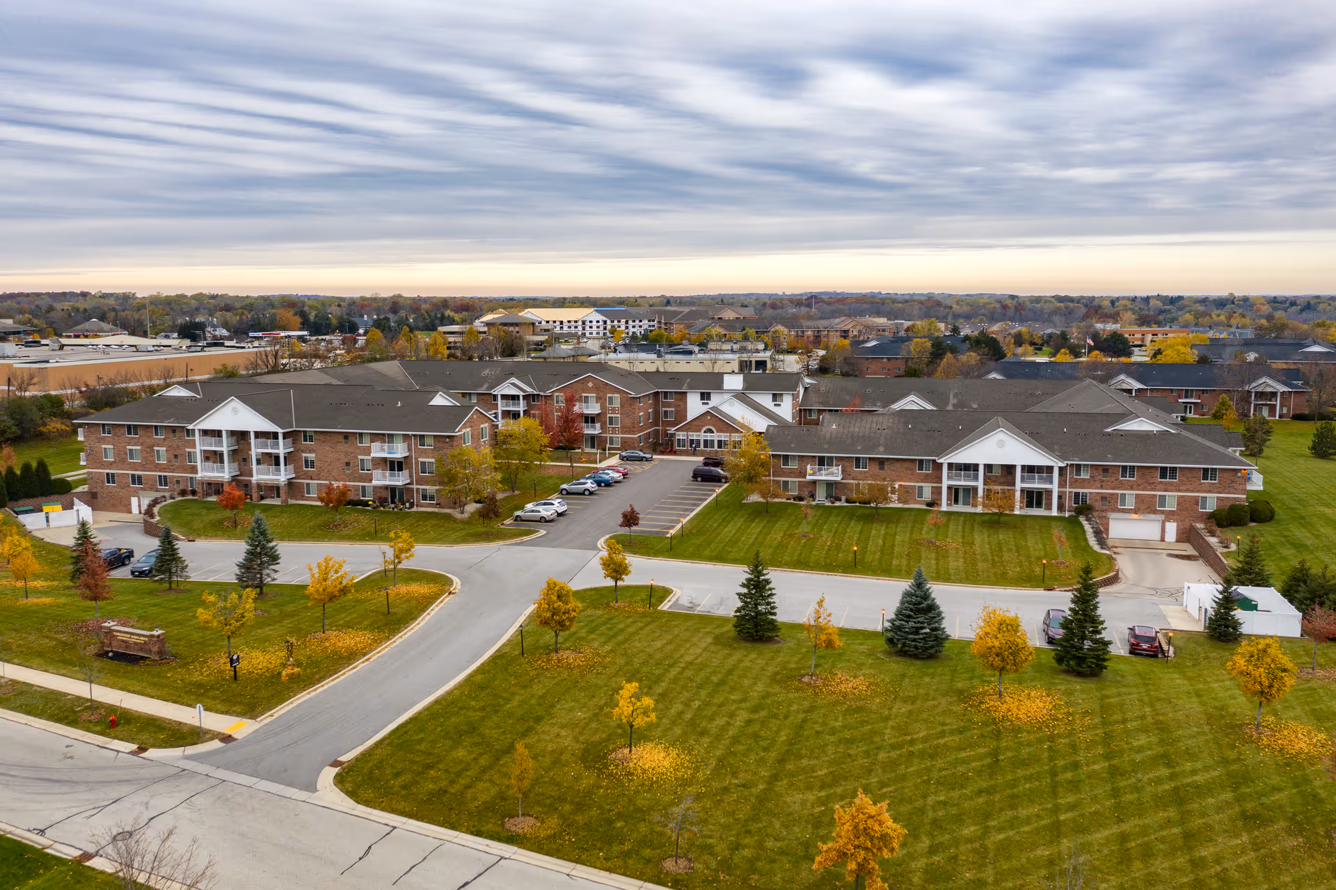 Aerial view of Parkwood Highlands Apartments & Townhomes 55+ complex showing multiple brick residential buildings with white balconies, surrounded by well-maintained green lawns, trees with autumn foliage, parking areas, and a cloudy sky overhead.