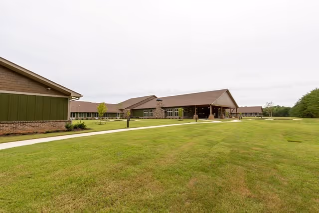 Wide view of a single-story assisted living facility with brown roofs and green siding, surrounded by a large grassy lawn and a paved walkway leading to the entrance under a covered porch.