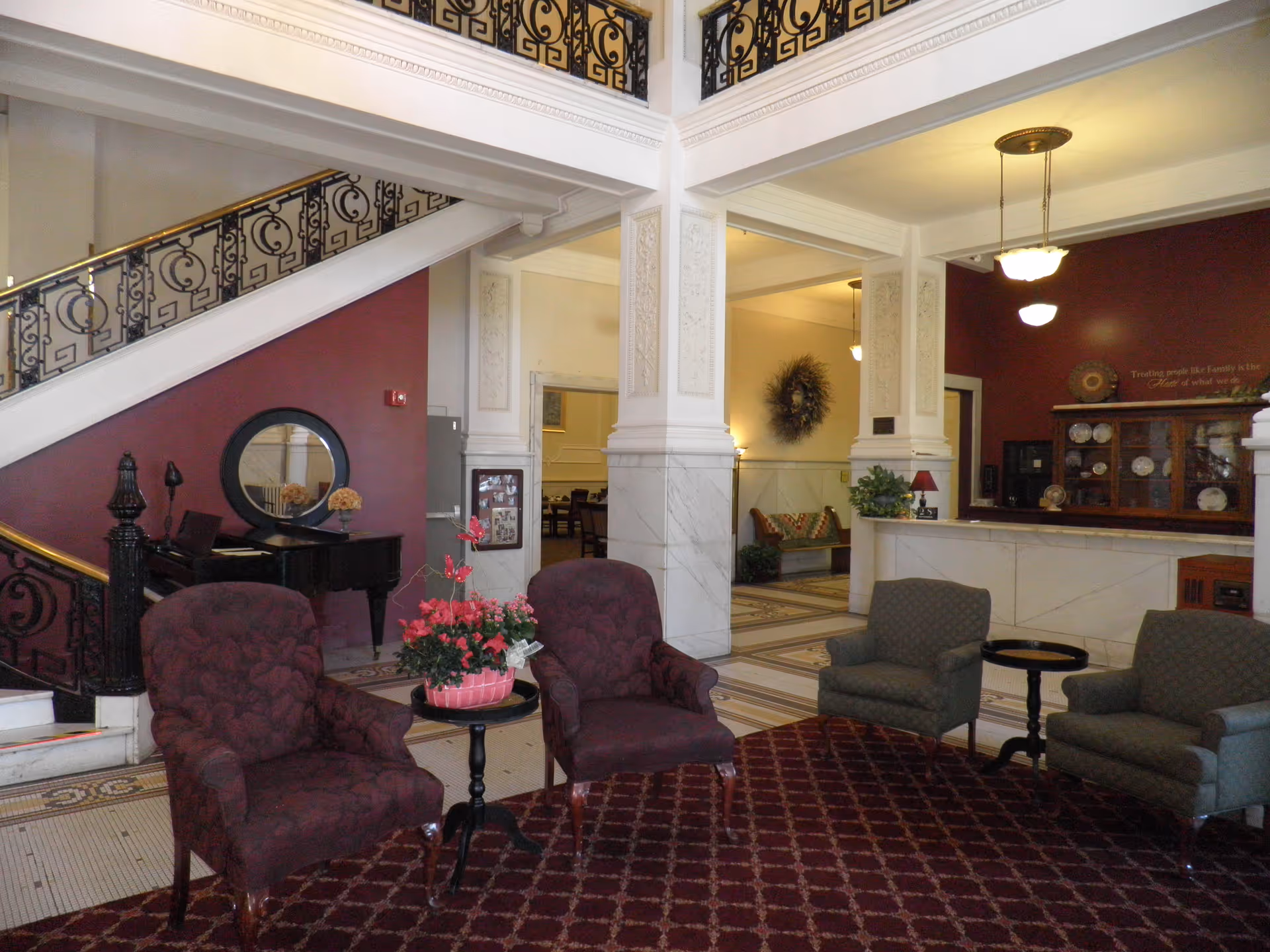 Interior view of a senior living facility lobby with a seating area featuring four upholstered chairs around small tables with flower arrangements. The space has a decorative staircase with ornate railings, marble columns, and a reception desk with display shelves and plates. Warm lighting fixtures hang from the ceiling, and a wreath decoration is visible in the background.