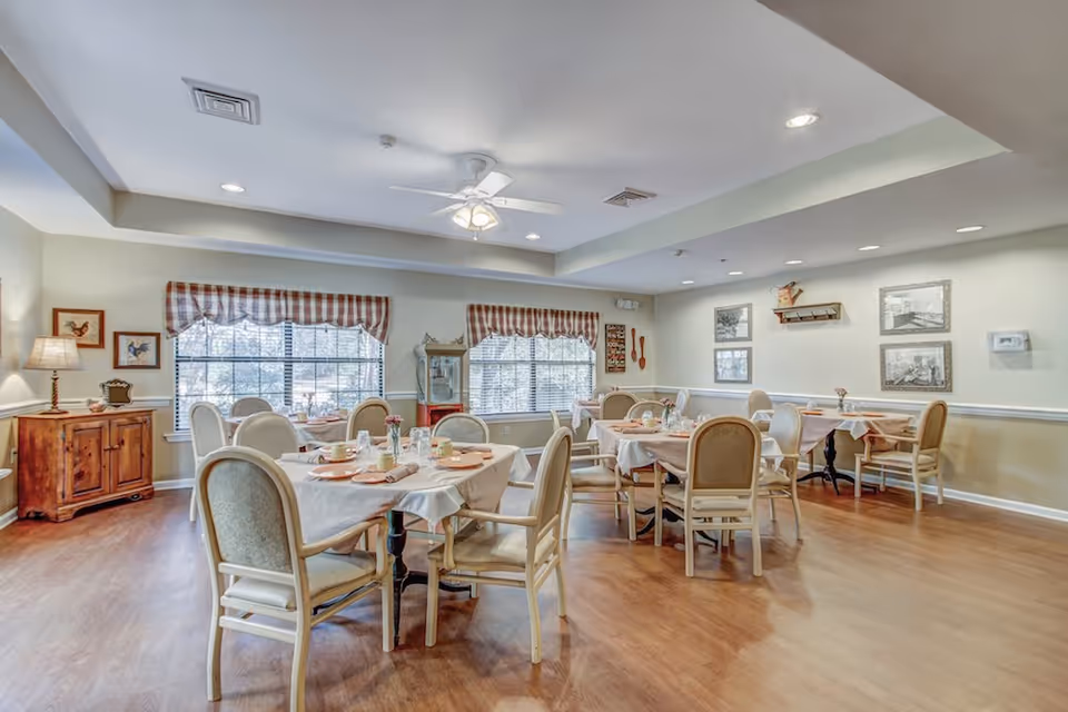 A bright dining room with several round tables covered with white tablecloths and set with plates, glasses, and napkins. The room has wooden flooring, beige walls with white trim, and large windows with checkered valances. There are cushioned chairs around the tables, framed pictures on the walls, a wooden cabinet with a lamp, and a ceiling fan with lights.