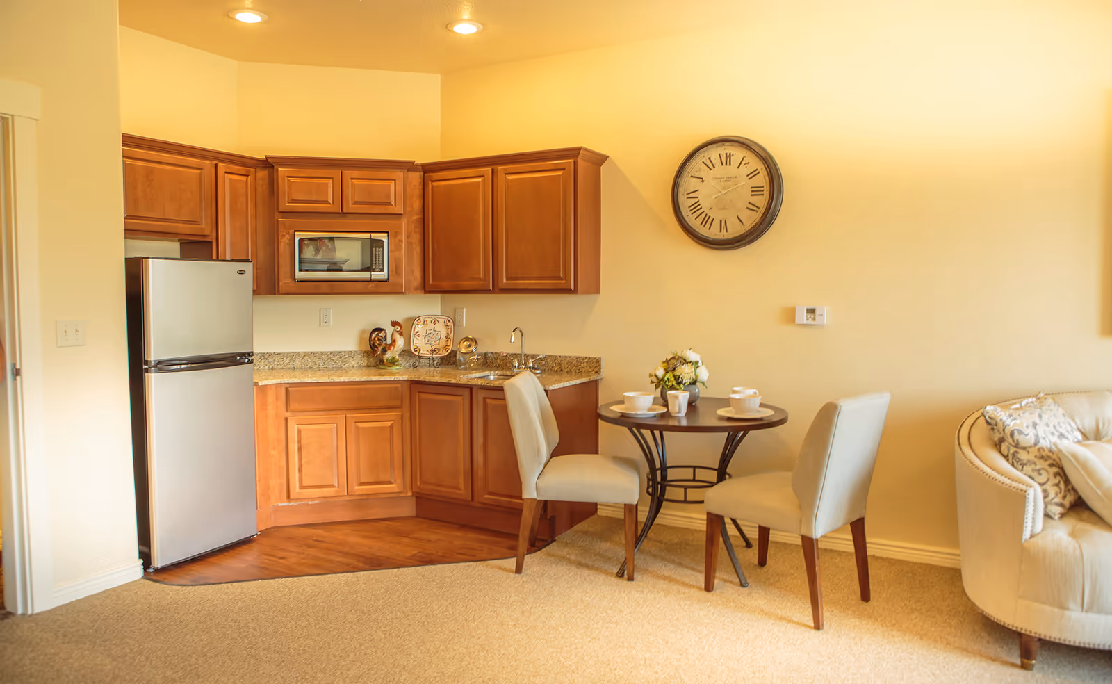 Small assisted living kitchenette with wooden cabinets, a stainless refrigerator, round dining table with two chairs, and a sofa.