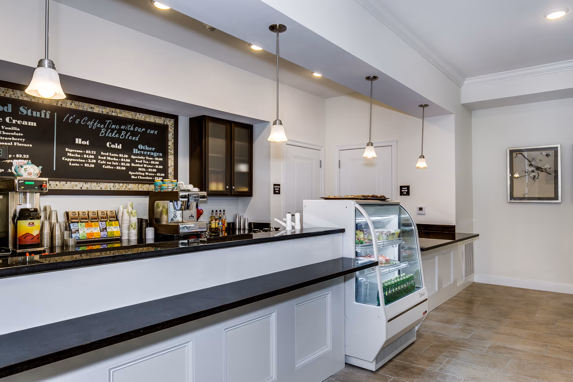 Bright indoor cafe-style serving counter with a menu board, coffee machines, pendant lights, and a refrigerated display case.