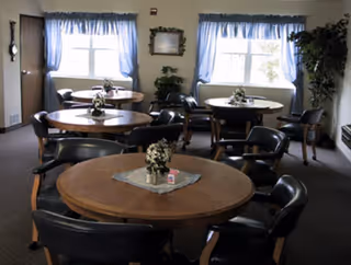 A dining room with round wooden tables and black chairs arranged around them. Each table has a small floral centerpiece and a lace doily. The room has two windows with blue curtains, a framed picture on the wall, and a potted plant in the corner.