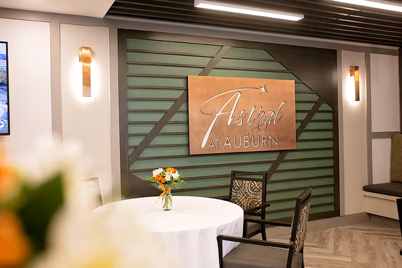 Round table with a small bouquet and chairs in a seating area beneath an "Astral at Auburn" sign on a decorative green wall.