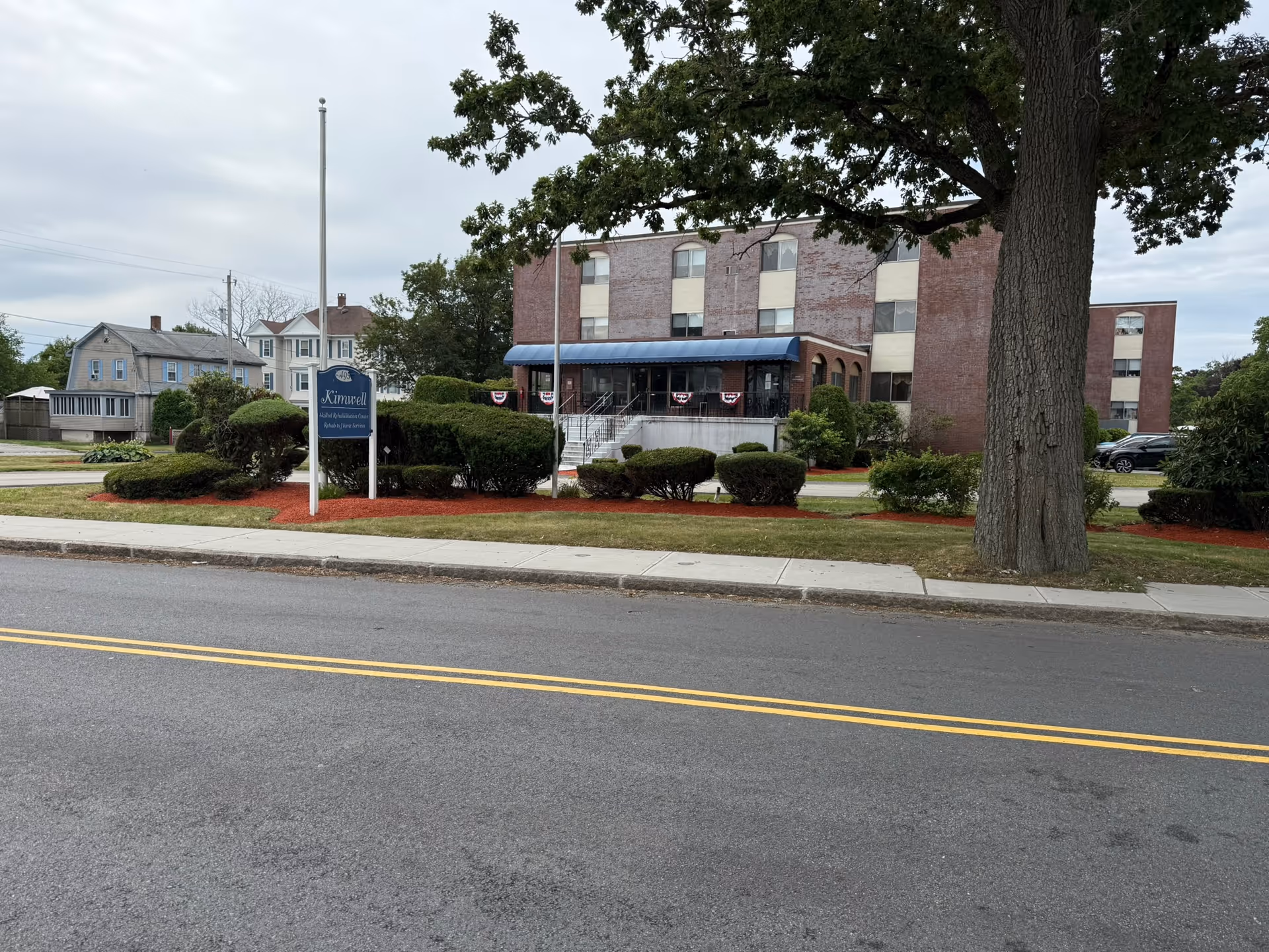 Exterior view of Kimwell Nursing and Rehabilitation building with a brick facade, a blue awning over the entrance, trimmed bushes, a large tree, and a sign on the lawn near the sidewalk. The sky is cloudy and there are houses visible in the background.