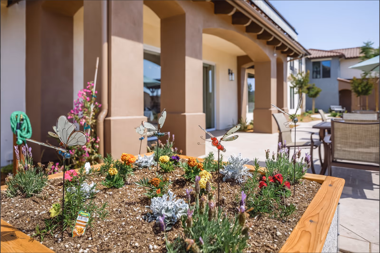 A raised garden bed with various colorful flowers and decorative metal insects in front of a beige building with arches. Patio furniture and umbrellas are visible in the background under a clear blue sky.