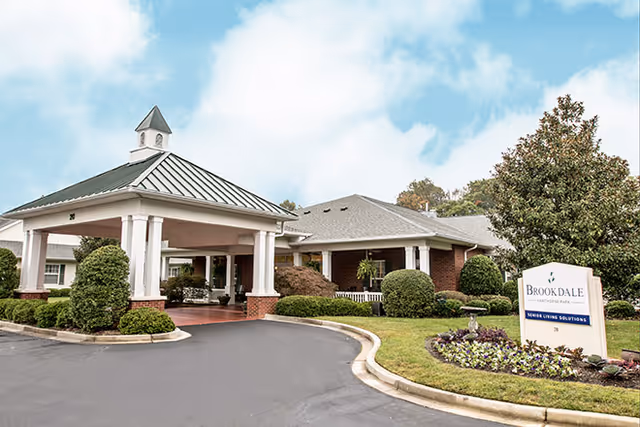 Exterior view of a senior living facility with a covered entrance supported by white columns, surrounded by well-maintained bushes and landscaping. A sign in front reads 'Brookdale Senior Living Solutions'. The sky is partly cloudy.